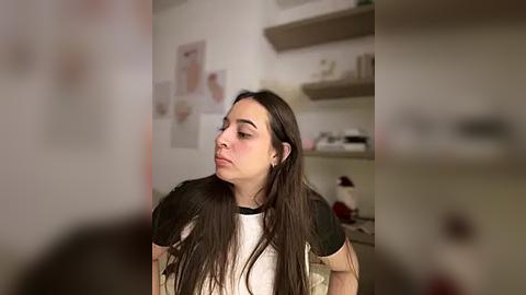 Media: Video of a young woman with long dark hair, wearing a black and white t-shirt, sitting indoors with shelves and anatomical posters in the background.