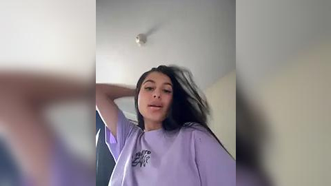 Media: A video of a young woman with long black hair, light skin, and a slender build, wearing a light purple t-shirt, captured from a slightly blurry, low angle.