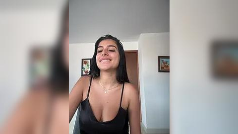 Media: Video of a smiling, light-skinned woman with long dark hair, wearing a black, low-cut spaghetti strap top, standing in a minimalist, white-walled room with framed art on the walls.