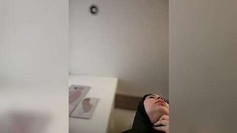 Media: A video of a woman with fair skin and dark hair, wearing a black hijab, lying on a white surface, with a medical chart and an anatomical illustration in the background.