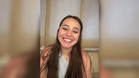 Media: Video of a smiling young woman with long brown hair, wearing a light pink cardigan over a white top, set against blurred kitchen cabinets in the background.