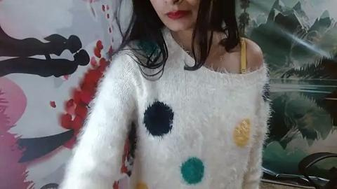 Media: Video of a woman with long black hair, wearing a white off-shoulder knit sweater with colorful polka dots, against a backdrop of abstract art featuring red and black splashes.