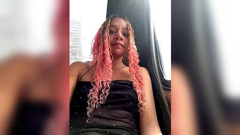 Media: Video of a young woman with long, pink curly dreadlocks and a black tank top, sitting in a car. Background includes blurred cityscape and car seat.