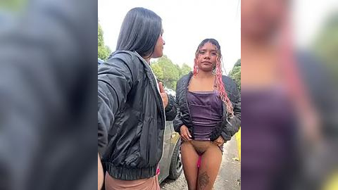 Media: Video of two young women, one with long black hair in a black jacket, the other with pink-dyed hair in a purple top, standing on a rural road.