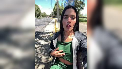 Media: Video of a woman with long dark hair, wearing a green top and gray jacket, sitting on a bench, making a surprised face, under a sunny sky with trees and a road in the background.