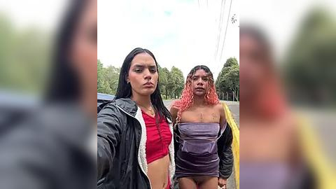 Media: Video of two young women with long hair, one in a red crop top and black jacket, the other in a purple strapless top, walking down a yellow-paved road. Background features blurred trees and an overcast sky.