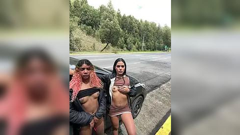 Media: Video of two young women in revealing outfits, standing beside a car on a rural road with a forest background. One has pink-dyed hair and a black jacket, while the other has long black hair and a brown crop top.