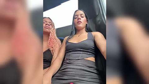 Media: Video of two young women with medium skin tones, one with braids and the other with straight hair, wearing matching gray tank tops and skirts, seated in a car with a blurred background.