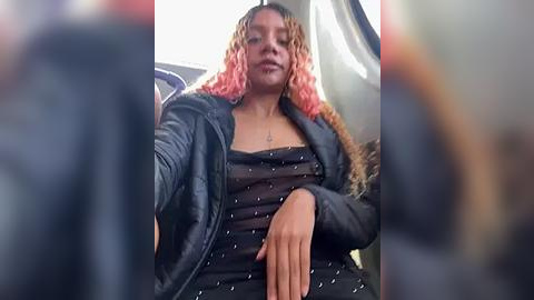 Media: Video of a young woman with medium brown skin, long curly pink and blonde hair, wearing a black leather jacket and a sheer black top with white polka dots, smiling slightly.