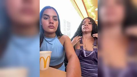 Media: Video: Two young women with medium skin tones, one in a light blue crop top and the other in a purple dress, sit in a fast-food restaurant with a blurred background.