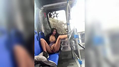 Media: Video of a woman with long black hair, sitting in a blue recliner chair on a bus, wearing a striped top and brown shorts, legs spread, looking out the window at a blurred landscape.