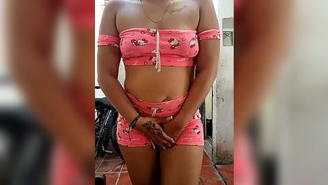 Media: Video of a woman in a pink, off-the-shoulder crop top and matching shorts, standing indoors with blurred background.