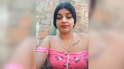 Media: Video of a young South Asian woman with short black hair, wearing a pink off-shoulder top with Hello Kitty pattern, sitting against a brick wall.