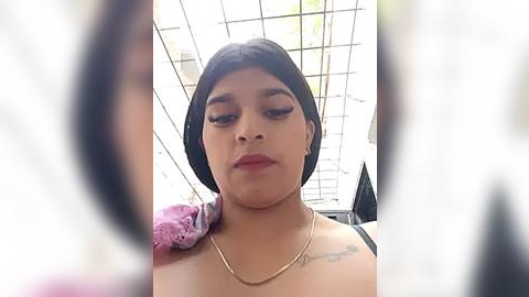 Media: Video of a person with medium skin tone, short black hair, and full makeup, including dark eyeliner and mascara, wearing a pink lace bra. They stand in a brightly lit room with a white ceiling and grid pattern.