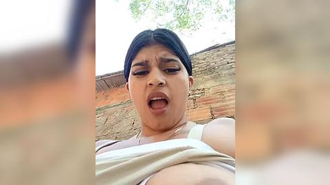 Media: A video of a Latina woman with dark hair, wearing a white tank top, exclaiming with her mouth open, against a rustic brick wall.