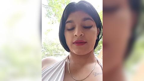 Media: Video of a young woman with medium skin tone, short black hair, and a confident expression, wearing a white off-shoulder top and a gold necklace. Background is blurred, showing green foliage.