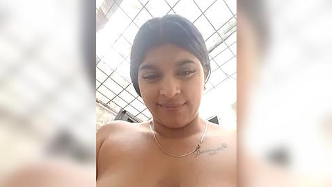 Media: A video of a young, light-skinned woman with medium-length black hair, smiling, wearing a thin necklace, taken indoors under a skylight. Her bare shoulders and upper chest are visible.
