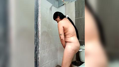 Media: Video of a naked, light-skinned woman with long black hair in a bathroom, standing with her back to the camera, looking down. The bathroom has white tiled walls and a toilet.