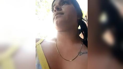 Media: Video of a young woman with medium skin tone, dark hair, and a yellow top, wearing a silver necklace, looking up with a thoughtful expression against a sunlit background.