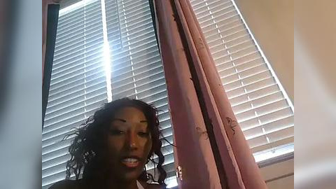 Media: Video of a young African-American woman with curly hair, standing in a room with beige walls, a window with white blinds, and pink curtains. She has a neutral expression, wearing a light-colored top.