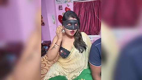 Media: A video of a South Asian woman with medium skin tone, wearing a yellow blouse, black mask, and green saree, adjusting her mask in a dimly lit room with pink walls and red curtains.