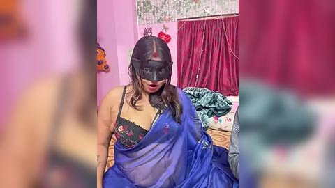 Media: Video of a South Asian woman with long, wavy hair, wearing a blue sari, black mask, and a black lace bra, sitting in a colorful bedroom with red curtains and decorative items.