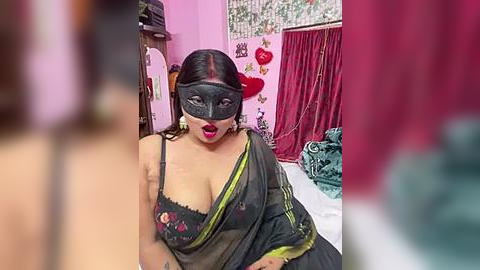 Media: Video of a woman in a black mask, dark green sari, and red lipstick, sitting on a bed in a pink room with red curtains and heart decorations.