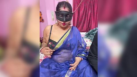 Media: Video of an Indian woman with medium skin tone, dressed in a blue saree, wearing a black masquerade mask, sitting on a bed with a red curtain in the background.