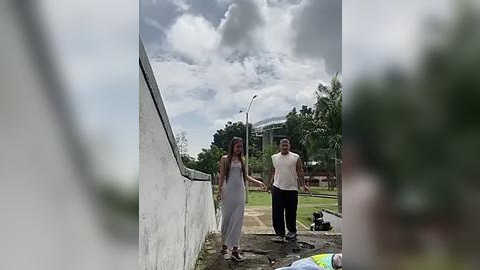 Media: Video of a young woman in a white dress and a man in a white tank top and black pants walking hand-in-hand along a concrete path beside a white wall in a suburban neighborhood.