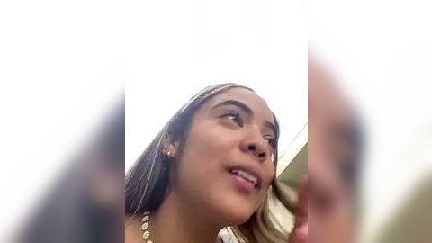 Media: Video of a young woman with medium brown skin, long straight brown hair, wearing a white dress, looking upward, with a blurred background.