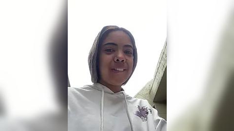 Media: Video of a young Black woman with a medium complexion and short hair, wearing a white hoodie with a floral design on the left chest, smiling against a blurred background.