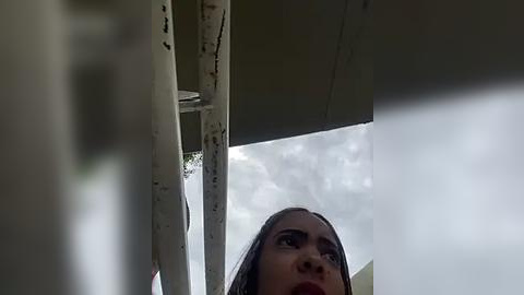 Media: A video of a woman with long dark hair, looking upwards, with a white metal structure and a cloudy sky in the background.