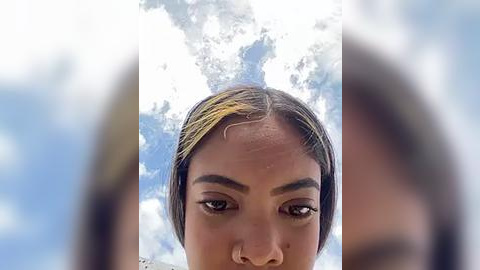 Media: Video of a woman with dark skin, short brown hair, and a prominent nose ring, set against a cloudy blue sky background.