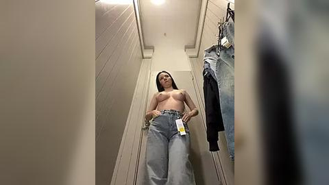 Media: Video of a topless woman with medium-sized breasts, wearing high-waisted jeans, standing in a narrow, beige hallway with clothes hanging on the right.