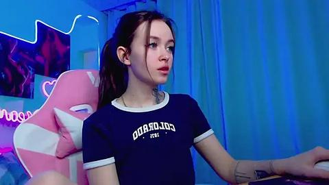 Media: Video of a young woman with fair skin and dark hair, wearing a navy blue \"Guardians of the Galaxy\" T-shirt, seated in a pink gaming chair, with a neon blue room in the background.