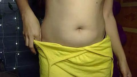 Media: Video of a fair-skinned person with a protruding belly, wearing yellow shorts, holding them open to reveal the navel. Background includes a dark curtain with string lights and a wooden surface.