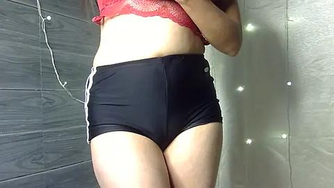 Media: Video of a fair-skinned woman in a red lace bra and black high-waisted shorts with white stripes, standing in a tiled bathroom with a string of white lights.