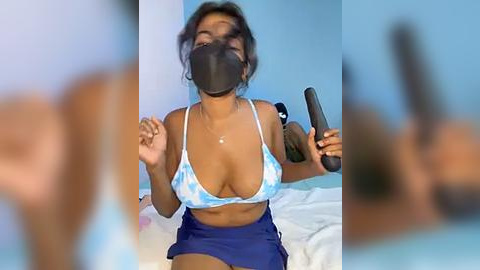 Media: Video of a dark-skinned woman with medium-sized breasts in a light blue tie-dye bikini top, dark hair, wearing a black face mask, holding a remote, seated on a bed in a blue room.