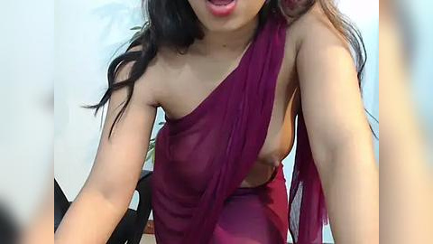 Media: Video of an Asian woman with long black hair, wearing a sheer, deep maroon wrap dress that exposes her left breast, smiling. Background features a blurred indoor setting with white walls and a hint of greenery.