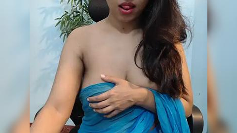 Media: Video of an Asian woman with long dark hair, wearing a blue strapless dress, covering her left breast with her hand, in a room with a potted plant in the background.