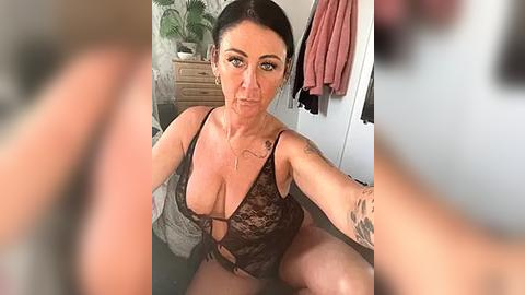 Media: Video of a middle-aged woman with fair skin and dark hair in a low-cut, black lace lingerie bodysuit, sitting on a bed in a cluttered, dimly lit room.