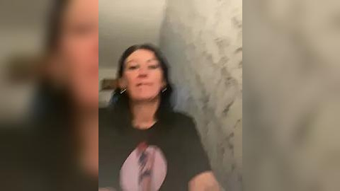 Media: Video of a woman with dark hair, wearing a black T-shirt with a pink graphic, standing in a dimly lit room with a white textured wall.