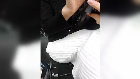 Media: Video of a woman with a large bust, wearing a tight, white, ribbed sweater, partially unbuttoned black cardigan, and holding a smartphone.