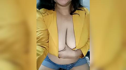 Media: A video of a woman with medium skin tone, wearing a mustard yellow blazer that's open to reveal her large breasts, and denim shorts.