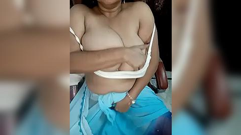 Media: Video of a topless, light-skinned woman with medium-sized breasts, wearing a blue skirt and holding a white cloth over her chest, seated on a wooden chair.