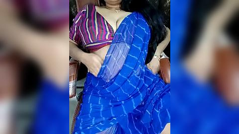 Media: Video of a woman in a blue saree with a white pattern, wearing a purple blouse with vertical stripes, sitting on a brown chair.