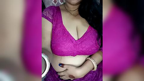 Media: Video of a South Asian woman with medium brown skin wearing a vibrant purple lace bralette, revealing ample cleavage, and a matching skirt. She's seated, her hands on her stomach, adorned with a gold bracelet. Background is blurred, focusing on her attire and jewelry.