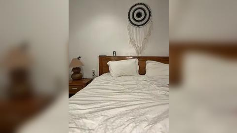 Media: Video of a minimalist bedroom featuring a neatly made bed with white linens, a wooden headboard, and a macram\u00e9 wall hanging above it. A nightstand with a lamp and a small shelf is on the left.