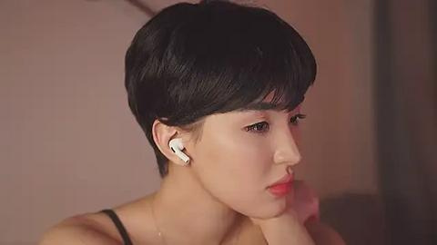 Media: Video of an East Asian woman with short, black, pixie-cut hair, wearing white earbuds and a black top, gazing thoughtfully at the right. Soft, warm lighting.