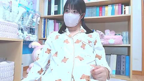 Media: Video of an East Asian woman with straight black hair, wearing a white pajama top adorned with panda patterns, and a white face mask. She is seated in a tidy, organized room with bookshelves, stuffed toys, and wicker baskets.